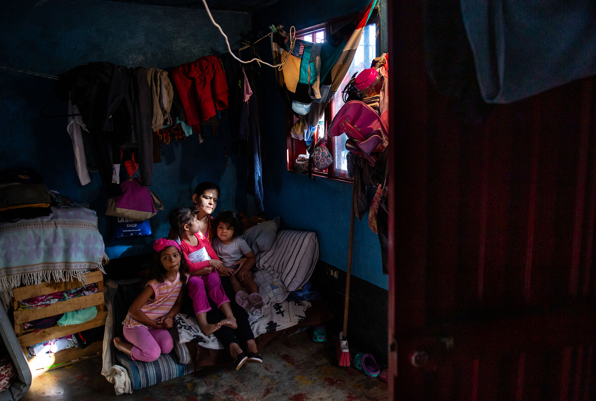 A mother holds her three children on a bed in their one room home as light hits her face