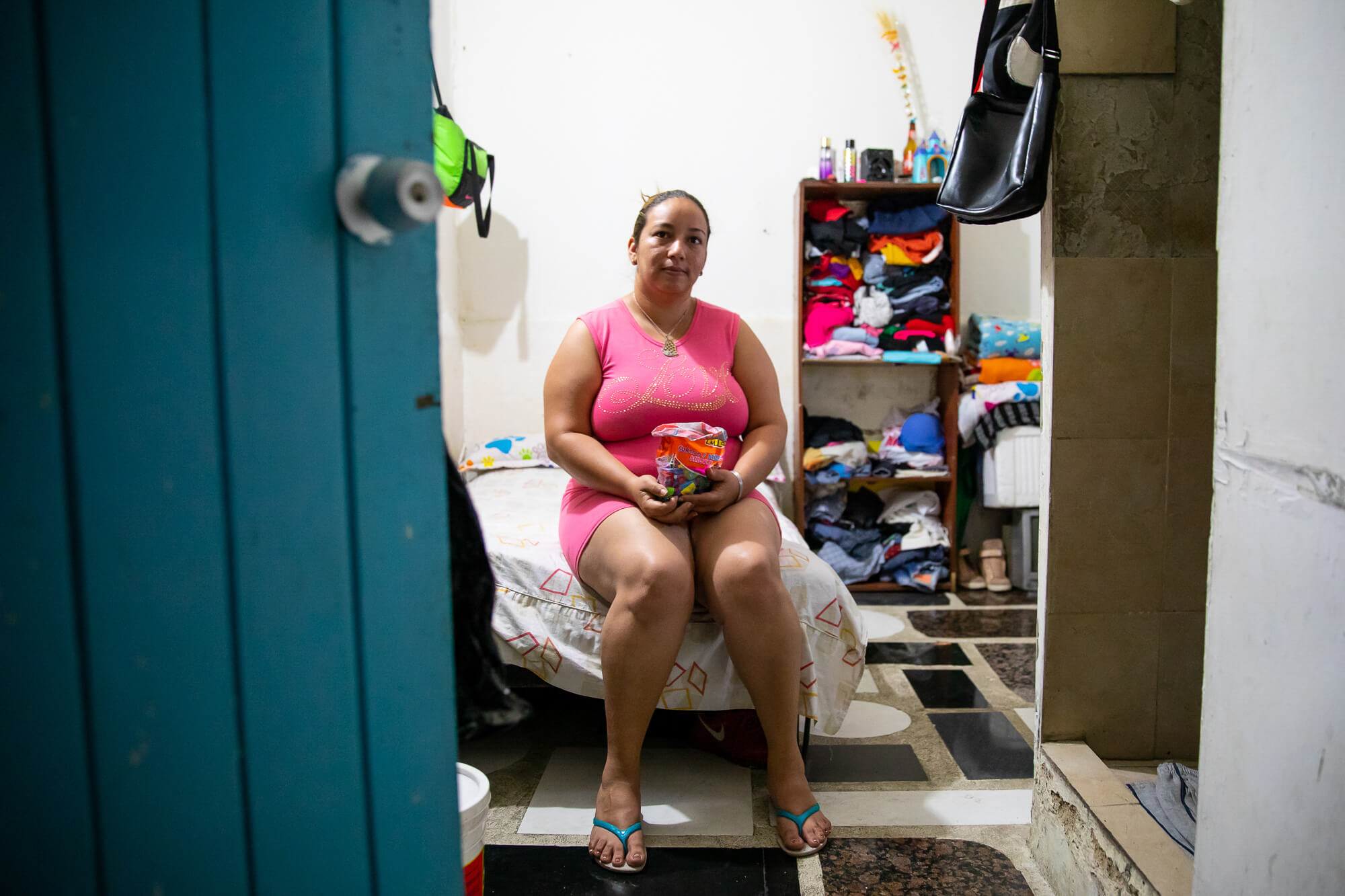 A woman wearing pink sits on her bed in a small rented room