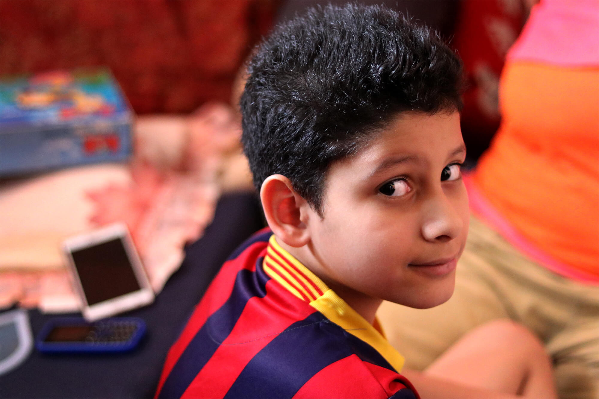 A young boy named Jeham Carlos looks over his shoulder at the camera. He wears a red and navy striped futbol jersey.