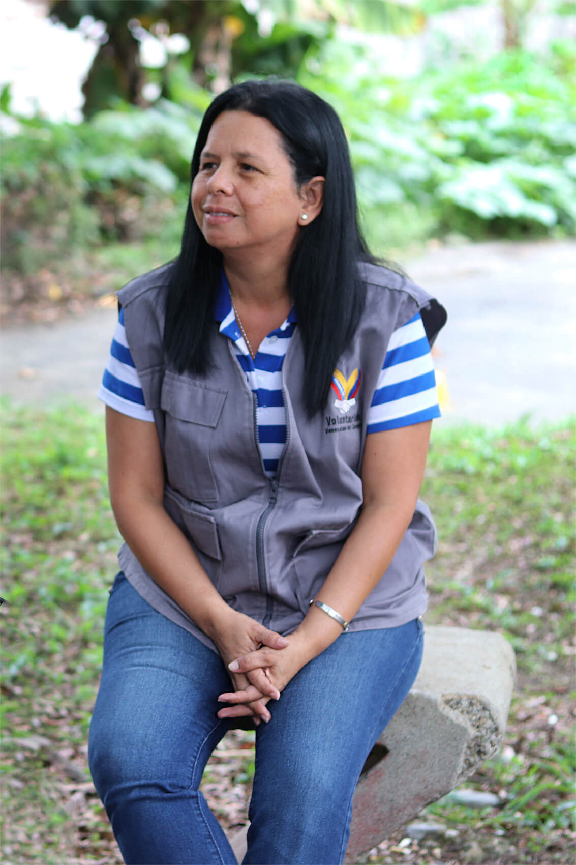 A woman named Lisbeth sits on a concrete bench outside. She wears blue jeans, a blue and white striped polo shirt, and a grey volunteer’s vest.