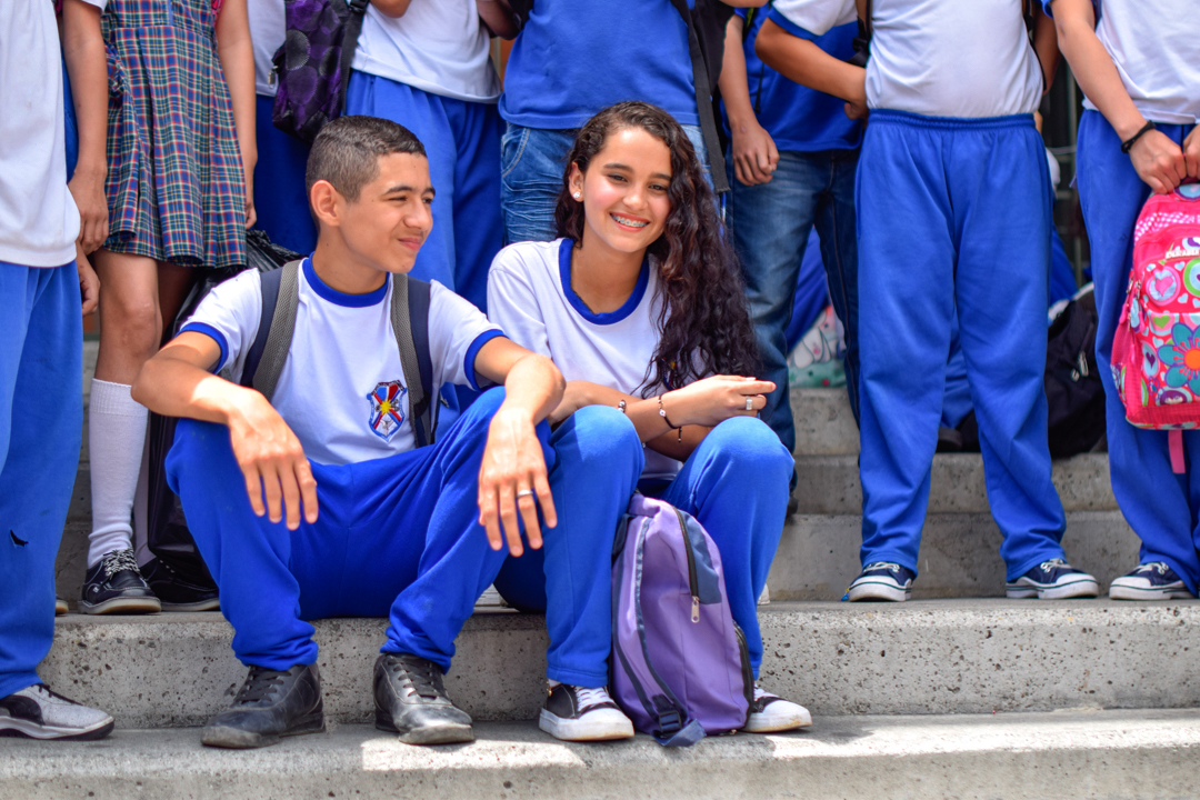 Two teenagers sit on the concrete steps of their school, smiling, surrounded by their peers. They wear blue and white uniforms.