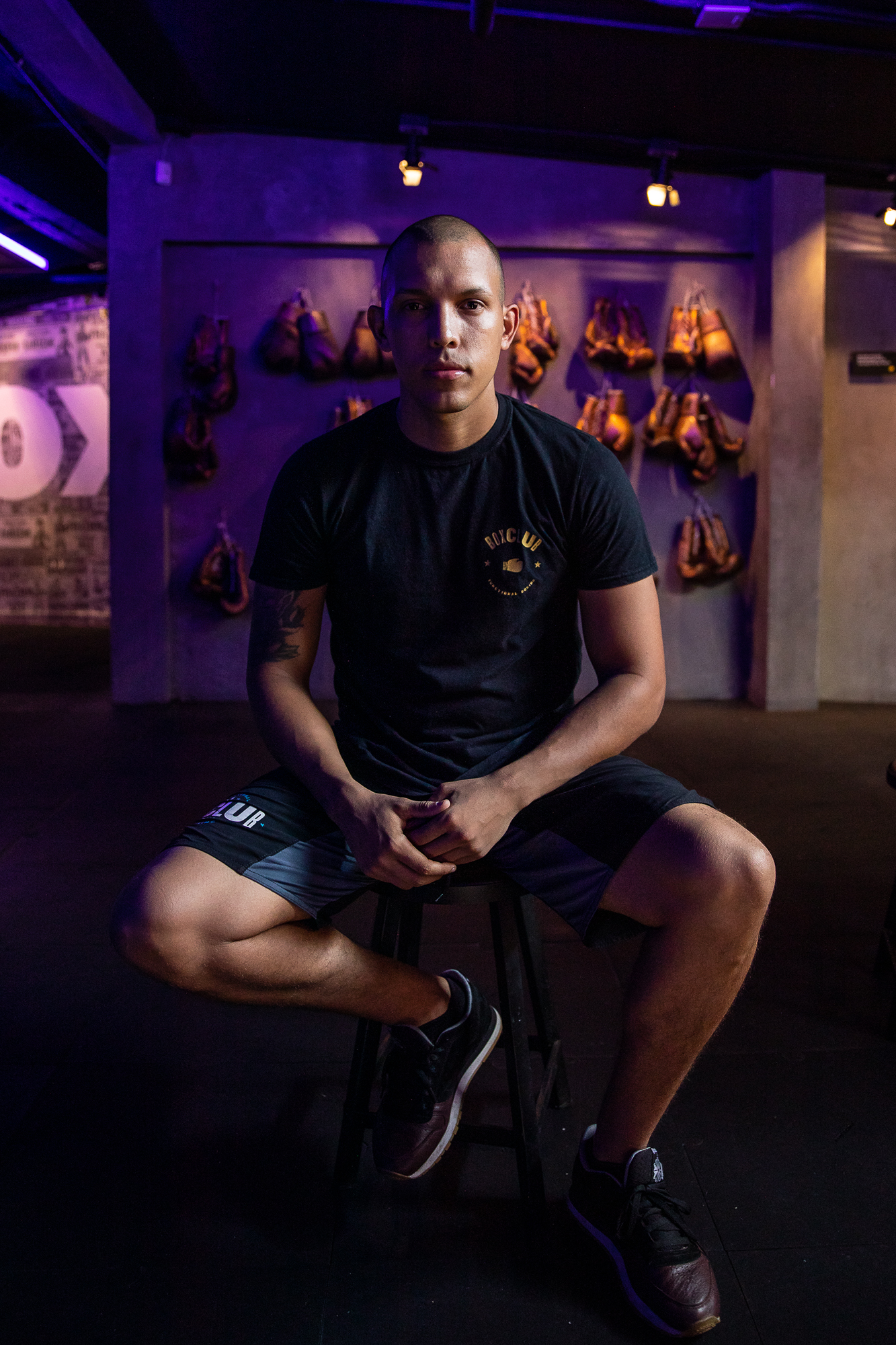 An athletic man in a t-shirt and shorts poses for a portrait inside of a boxing and fitness gym while sitting on a stool