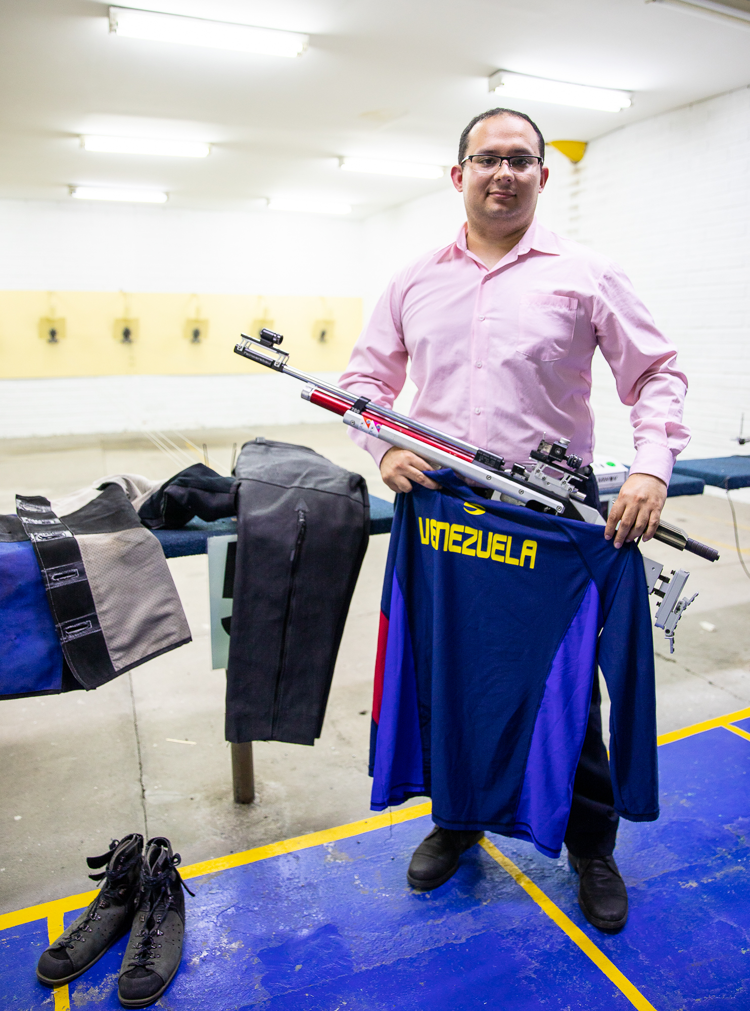 A man posing for a portrait in a button-up shirt holds an olympic shooting rifle while holding a Venezuela jersey inside of a shooting range