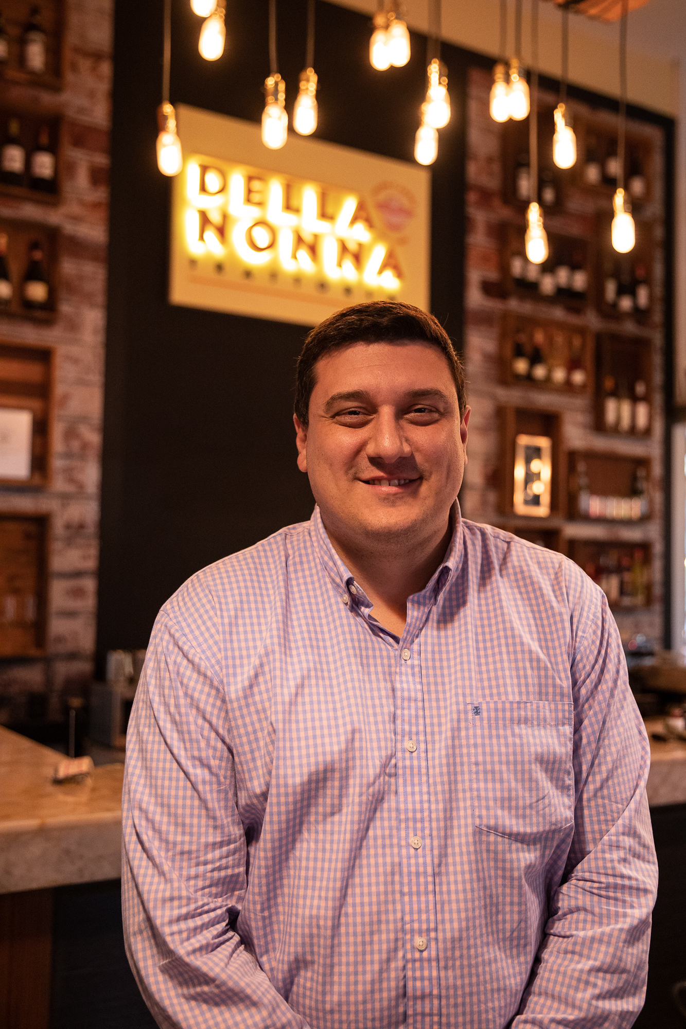 A man poses for a portrait in a button-up shirt inside of a restaurant while standing in front of a lighted sign that says 'Della Nonna'.