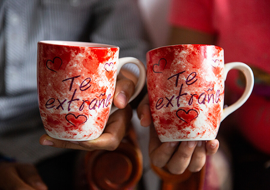 Two arms extended hold up matching mugs that read “Te extraño”